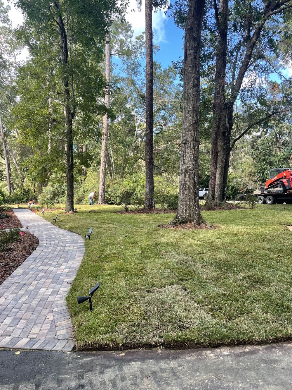 Lush green lawn with a pathway and trees, workers tending to landscaping in the background.