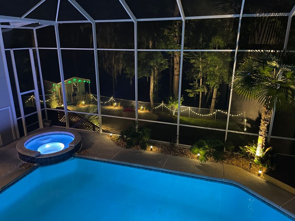 Outdoor pool and hot tub at night with illuminated garden and trees in the background.