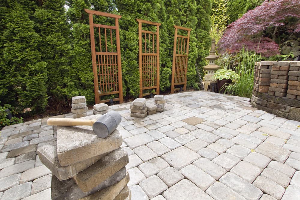 Paved garden patio with stone blocks and wooden trellises surrounded by greenery.